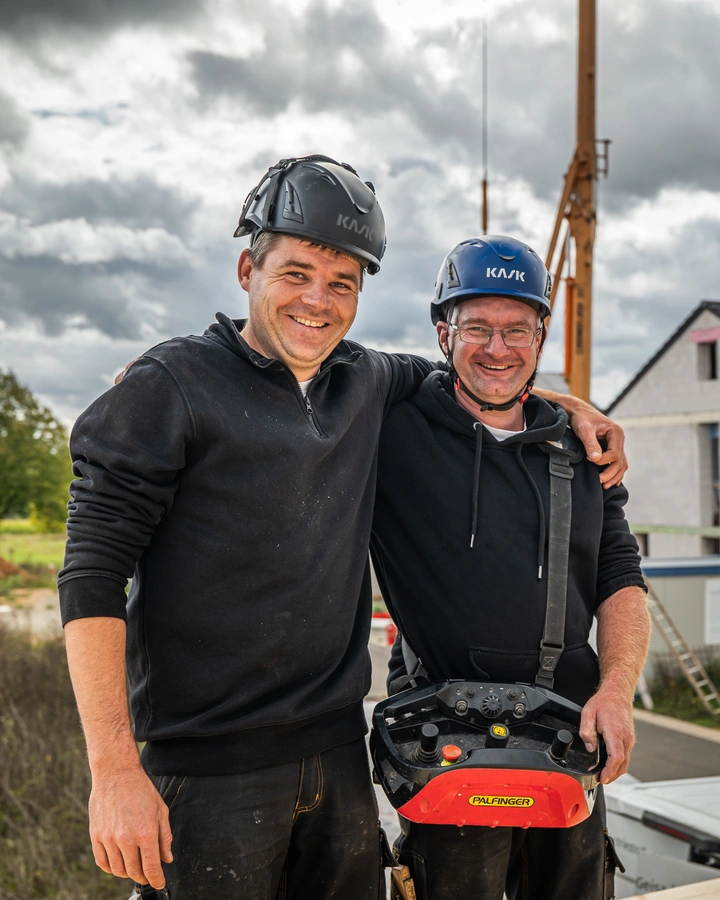 Zwei Handwerker mit Helmen stehen Arm in Arm auf einer Baustelle, dahinter ein Rohbau und ein Kran