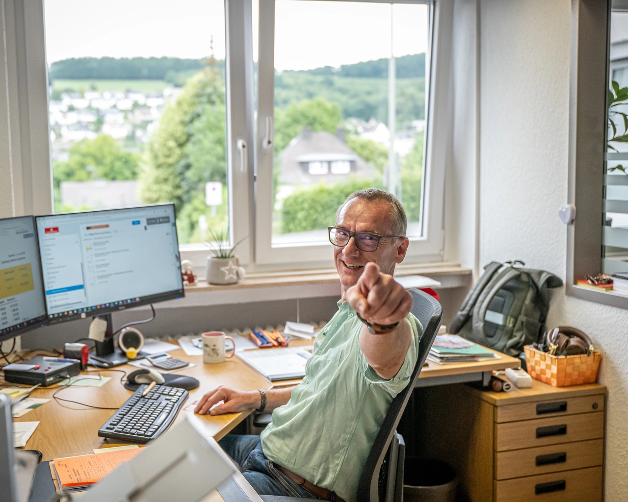 Mann mit Brille sitzt im Büro vor zwei Bildschirmen und zeigt lachend in die Kamera