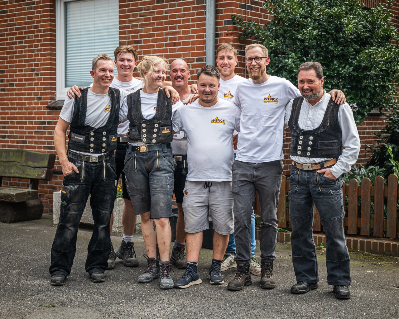 Acht Zimmerer in Zunftweste und weißen Shirts stehen lachend Arm in Arm vor einem Backsteinhaus