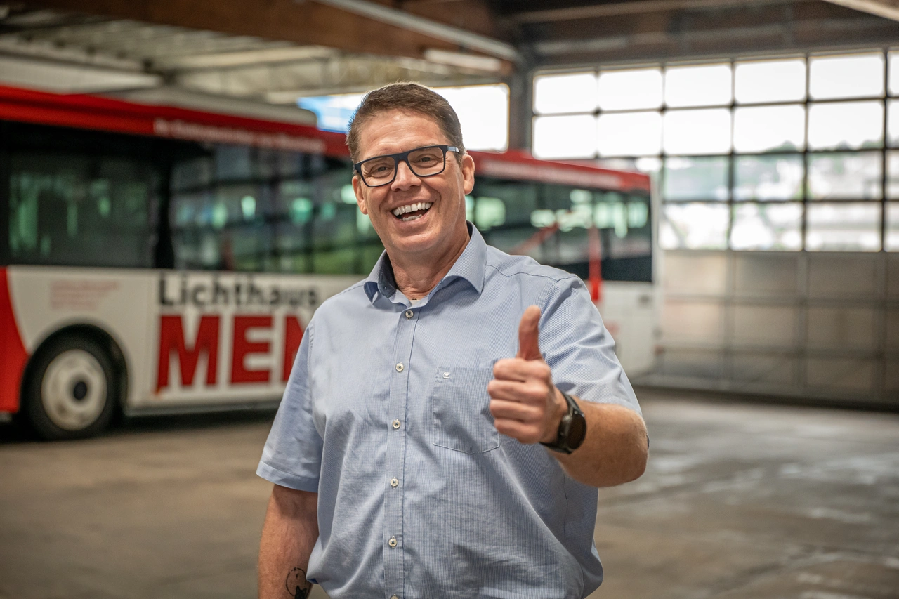 Ein Mitarbeiter lacht und zeigt den Daumen nach oben, hinter ihm steht ein Bus in der Halle