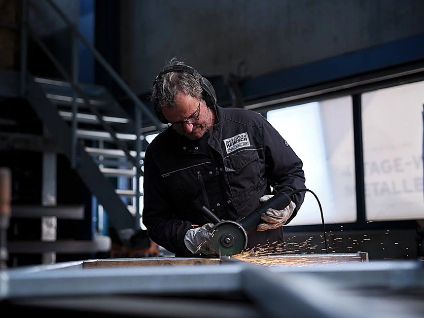 Metallbauer trennt mit dem Winkelschleifer ein Stahlprofil, Funken fliegen über die Werkbank, er trägt Gehörschutz und Schutzbrille