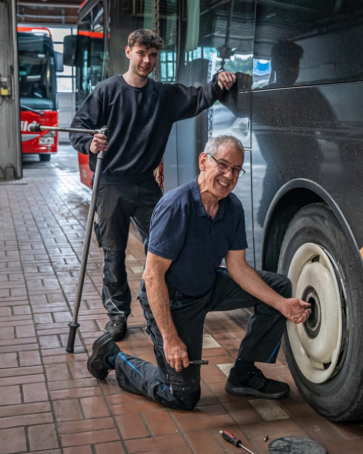 Älterer Mechaniker kniet am Rad eines Reisebusses, ein junger Kollege steht mit Werkzeug daneben