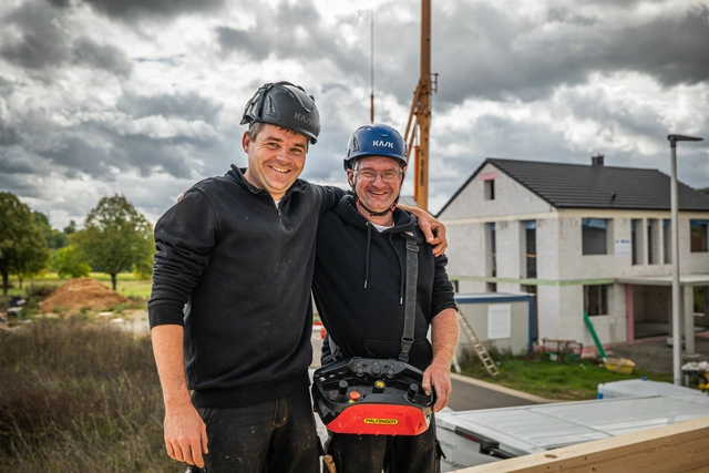 Zwei Kollegen in Arbeitskleidung und Schutzhelm stehen Arm in Arm auf einem Rohbau, einer trägt eine Kranfernbedienung vor dem Bauch