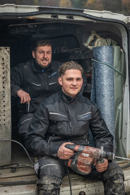 Zwei Handwerker in verschmutzter Arbeitskleidung sitzen auf der Ladekante eines Transporters, der vordere hält einen Bohrhammer