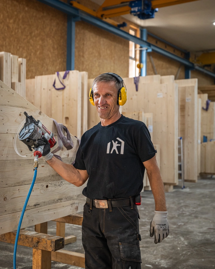 Zimmerer mit Gehörschutz hält ein Druckluftwerkzeug in einer Halle voller Holzelemente